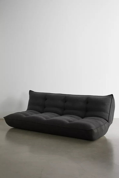 The Greta XL Sleeper Sofa, with its modern black tufted design, sits against a plain white wall on a polished concrete floor. Its minimalist style features a low back and no armrests, offering a sleek and contemporary appearance.