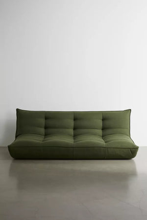 The Greta XL Sleeper Sofa, featuring a modern minimalist design with a tufted backrest, stands against a plain white wall on a smooth concrete floor. This green sofa has a low profile and no armrests, offering a sleek and contemporary appearance.