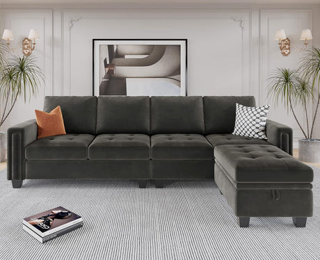 A modern living room showcases the Belffin Reversible Sectional Convertible Storage sofa in dark green, accented by cozy cushions. The decor includes two tall plants, wall sconces, and a large framed abstract artwork. On the floor lies a soft striped rug with a book resting on it.