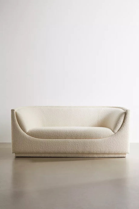 The Aria Sofa, with its minimalist cream tone and soft, curved design, is showcased against a plain white background on a smooth floor. It boasts textured fabric and exudes a modern aesthetic.