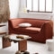 A modern living room features the Aria Sofa in a rust color, accompanied by a glass coffee table with wooden legs, a woven wall hanging, and a textured red rug. Sunlight streams through a large window, adding warmth to the minimalist decor. Decorative items are elegantly arranged on the table.