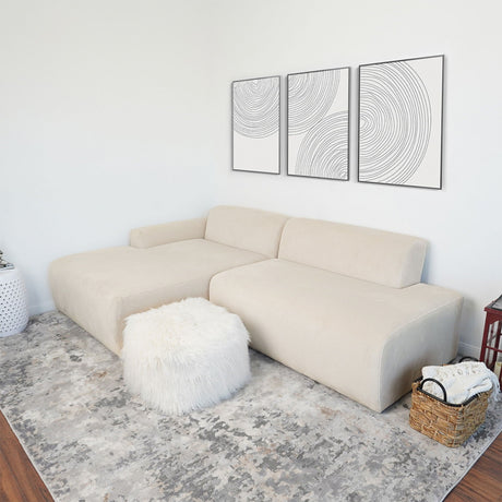 A minimalist living room features the Mar - High-Quality Sectional Sofa in beige, a fluffy white ottoman, and a gray patterned rug. Three abstract line art pieces adorn the wall, while a woven basket and white planter add decorative touches.