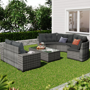 The outdoor patio features an 8 Pieces Outdoor Wicker Round Sofa Set, with a curved sofa and two chairs with dark cushions. A glass-top coffee table completes the set, all resting on grass. The area is framed by green hedges against a white house wall.