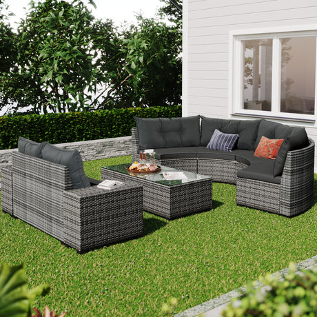 The outdoor patio features an 8 Pieces Outdoor Wicker Round Sofa Set, with a curved sofa and two chairs with dark cushions. A glass-top coffee table completes the set, all resting on grass. The area is framed by green hedges against a white house wall.