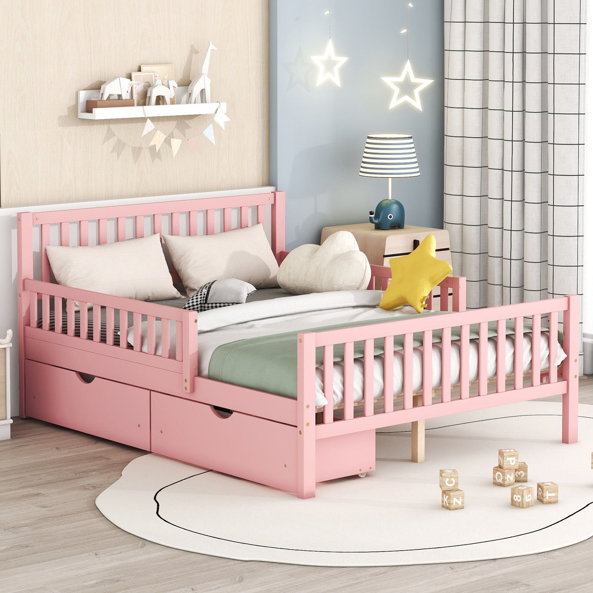 A children's bedroom has a pink platform bed with side guardrails and two storage drawers, white bedding, stuffed toys including a star-shaped pillow, hanging star lights, a table lamp, a bookshelf filled with books, and wooden blocks scattered on a round rug.