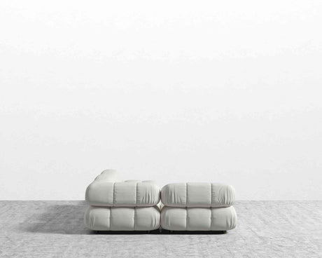 The Belia Modular Sectional, featuring tufted cushions in a light, soft fabric, is placed on a gray carpet against a white wall. The view from the back highlights its simple, modern design.