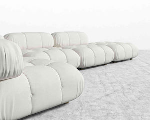 The Belia Modular Sectional in light gray upholstery is arranged on a matching light gray carpet. It features soft, rounded cushions with a tufted design, offering a modern and cozy look against the backdrop of a plain white wall.