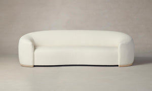 The Chelsea Sofa in Italian Bouclé White, featuring a modern and minimalist curved design with a plush texture, stands against a neutral background. Its rounded arms and low profile give it a sleek, contemporary look.