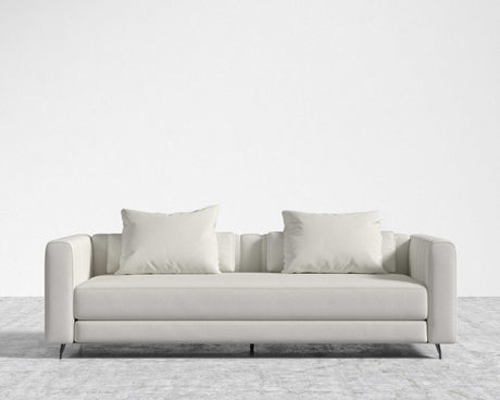 The Berlin Sleeper Sofa, featuring a modern and sleek minimalist design with smooth lines and subtle tufting on the arms, is placed against a plain white wall on a textured gray carpet. This light gray sofa comes with two matching square cushions.
