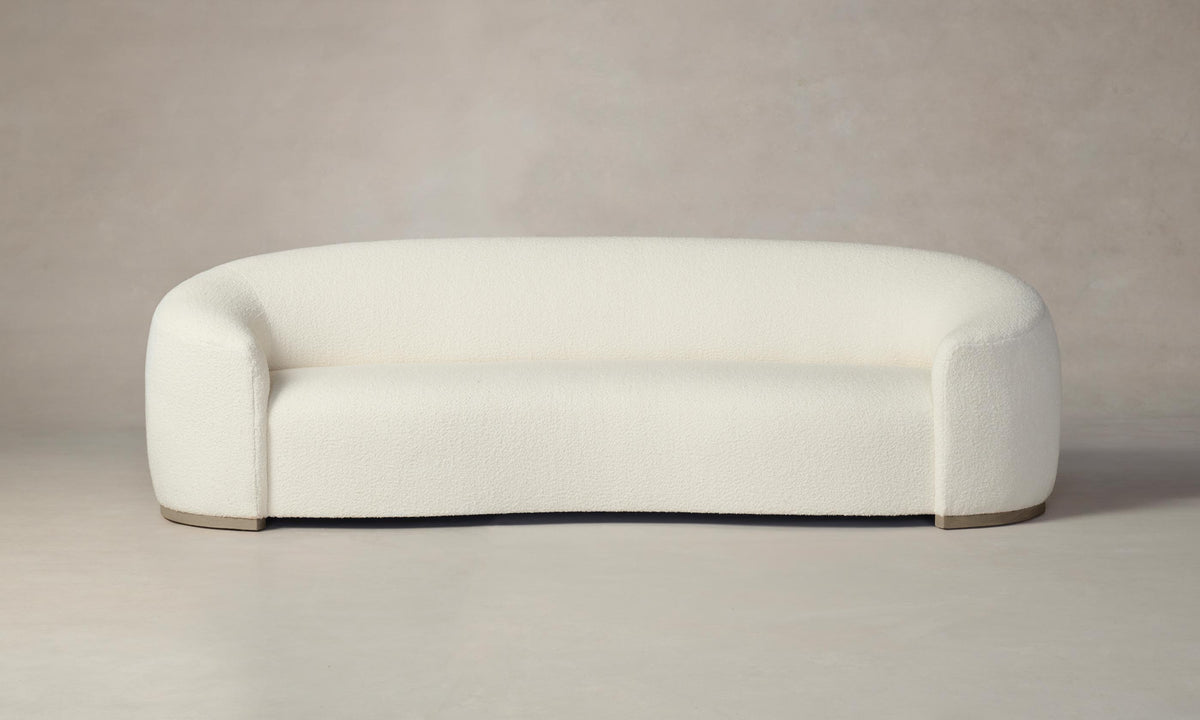 The Chelsea Sofa in Italian Bouclé White features a modern, minimalist aesthetic with its cream-colored, rounded design and smooth upholstery. Set against a neutral background, this sofa presents a clean and seamless appearance complemented by its slightly elevated base.