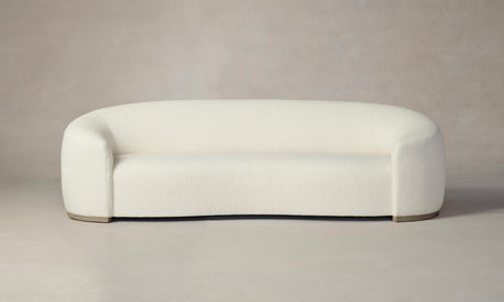 The Chelsea Sofa in Italian Bouclé White features a modern, minimalist aesthetic with its cream-colored, rounded design and smooth upholstery. Set against a neutral background, this sofa presents a clean and seamless appearance complemented by its slightly elevated base.