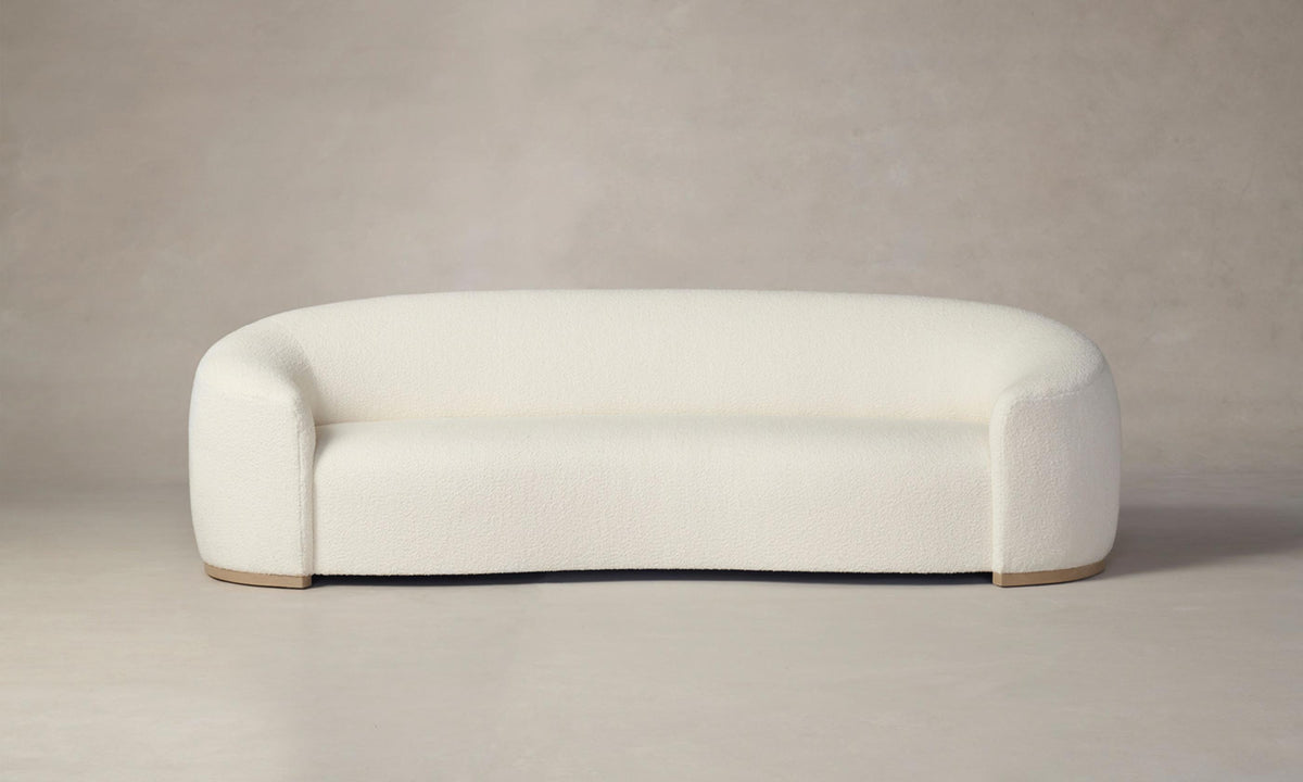 The Chelsea Sofa in Italian Bouclé White, featuring a modern and minimalist design with rounded curves, is displayed against a neutral gray background. Its plush upholstery and wooden base bring an elegant simplicity to the piece.