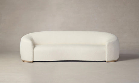 The Chelsea Sofa in Italian Bouclé White, featuring a modern and minimalist design with rounded curves, is displayed against a neutral gray background. Its plush upholstery and wooden base bring an elegant simplicity to the piece.