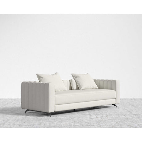 The Berlin Sleeper Sofa, with its modern light gray upholstery and vertically tufted sides and arms, is elegantly positioned on a textured gray rug. It includes two matching throw pillows and stands on sleek black legs, all set against a plain white background.