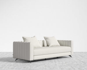 A Berlin Sleeper Sofa, featuring a modern and sleek ribbed design in light gray, sits on a matching light gray carpet. Two complimentary cushions enhance the minimalist and elegant setting against the plain white wall backdrop.