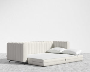 The Berlin Sleeper Sofa, featuring a modern and minimalist design in beige with a quilted pattern, is opened into a bed. Two white pillows rest on the mattress in a bright room adorned with a neutral-toned carpet and plain white walls.