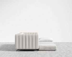 The Berlin Sleeper Sofa, featuring a minimalist and modern design in light gray with cushioned seating, is arranged against a plain white wall. This low-profile sofa includes a segmented backrest and is set on flooring covered in a light-colored textured carpet.