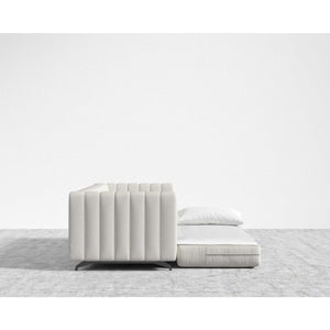 The Berlin Sleeper Sofa, in a contemporary light gray design, partially transforms into a flatbed and features a sleek pillow on top. It is set against a minimalist white wall and rests atop a gray carpeted floor.
