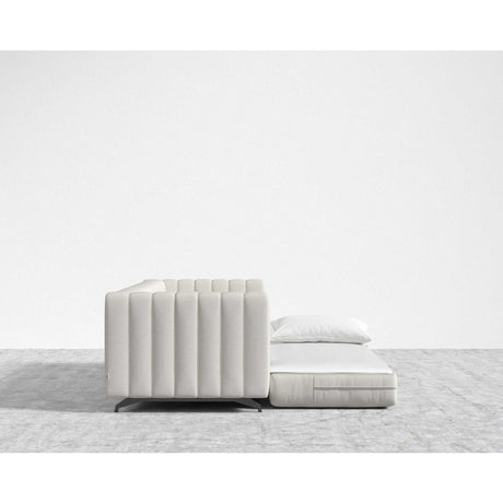 The Berlin Sleeper Sofa, in a contemporary light gray design, partially transforms into a flatbed and features a sleek pillow on top. It is set against a minimalist white wall and rests atop a gray carpeted floor.