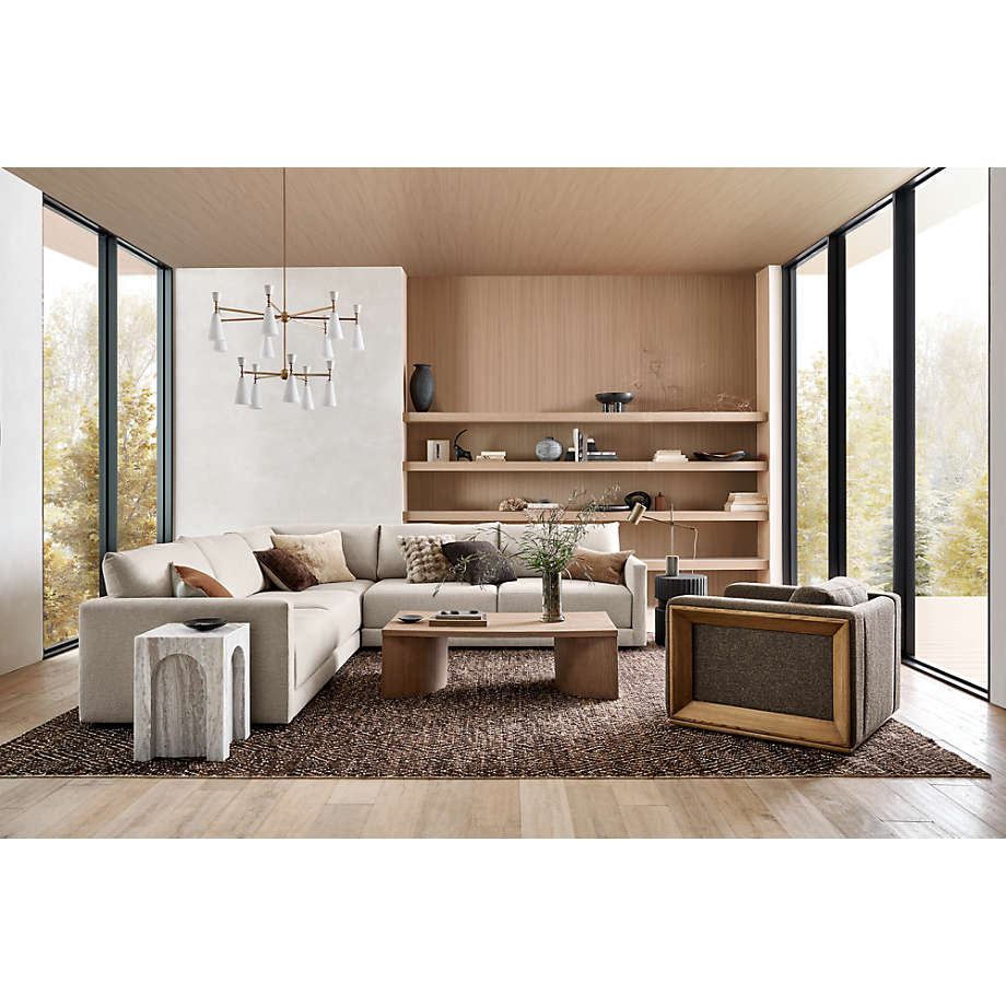 A modern living room with large windows showcases the Gather Sofa 89", a beige sectional complemented by a wooden coffee table and a textured area rug. Decor items are displayed on shelves against the wooden back wall. Pendant lights illuminate the space as natural light pours in through the windows.