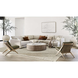 A modern living room features a neutral Gather Sofa 89" arranged in an L-shape and adorned with brown and white pillows. A round wooden coffee table rests on a textured rug, flanked by two wooden lounge chairs. Large potted plants and minimalist wall art complete the decor.