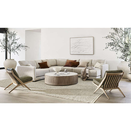 A modern living room features a neutral Gather Sofa 89" arranged in an L-shape and adorned with brown and white pillows. A round wooden coffee table rests on a textured rug, flanked by two wooden lounge chairs. Large potted plants and minimalist wall art complete the decor.