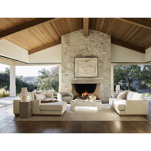 A spacious living room with a high wooden ceiling and large stone fireplace features two Tidal Apartment 83" Sofas and armchairs arranged around a geometric coffee table. Large windows provide views of the trees, filling the space with natural light. Above the fireplace, a framed abstract painting is displayed.