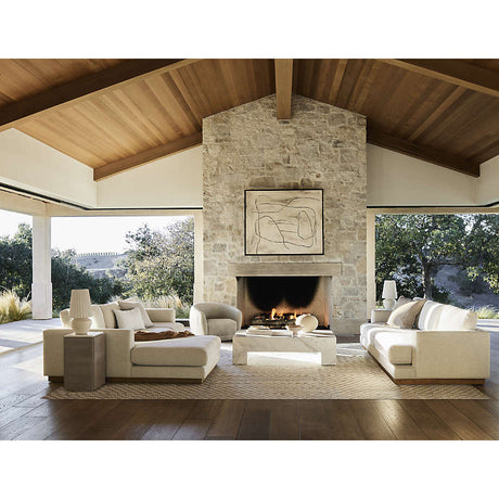A spacious living room with a high wooden ceiling and large stone fireplace features two Tidal Apartment 83" Sofas and armchairs arranged around a geometric coffee table. Large windows provide views of the trees, filling the space with natural light. Above the fireplace, a framed abstract painting is displayed.