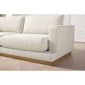 The Tidal Apartment 83" Sofa, a beige plush piece featuring large cushions and set on a wooden base, is placed on a light wood floor. A hint of framed artwork can be seen in the background.