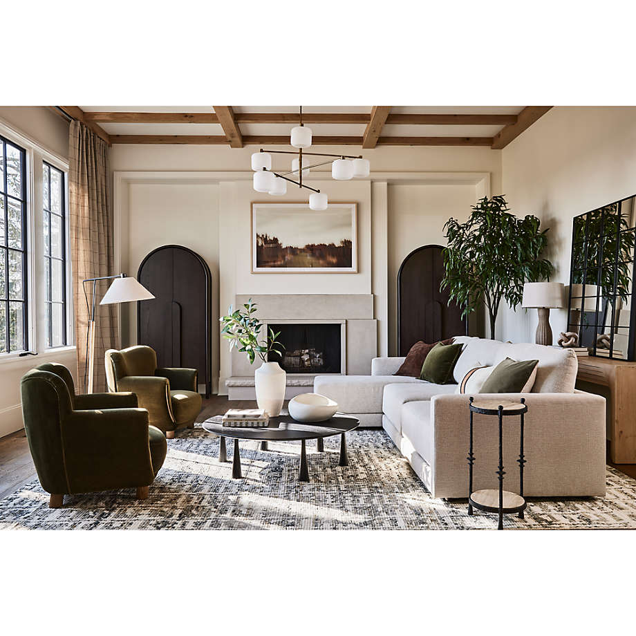 A modern living room showcasing a beige Gather Sofa 89" accented with green pillows, two green armchairs, and a round coffee table. A geometric chandelier is suspended from the wooden ceiling. The room also features a fireplace, large windows, and a potted plant.