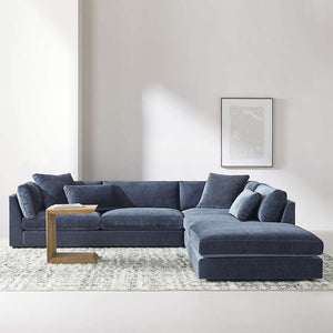 A modern living room showcasing the Monterey Modular 3 Piece L Shaped Sectional Sofa in dark blue, adorned with a variety of cushions. The space also includes a small wooden side table and a light-colored abstract painting on the wall, complemented by a neutral rug and light grey walls.