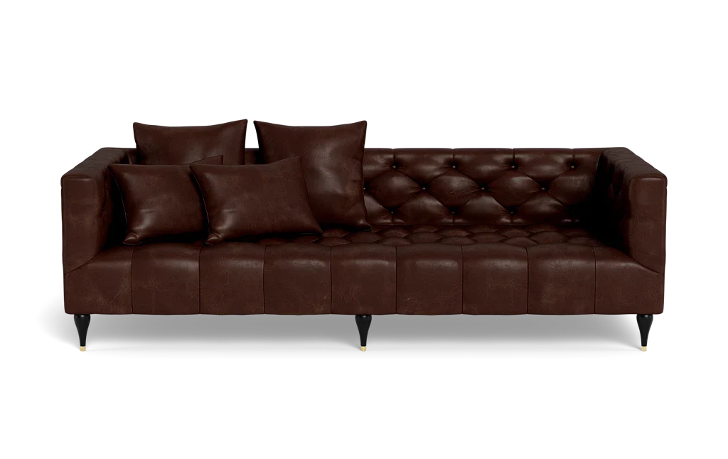The Ms. Chesterfield Leather Custom Sofa is a dark brown tufted leather piece featuring three seat cushions and four matching throw pillows. It boasts a classic design with black tapered legs and gold tips, set against a black background.