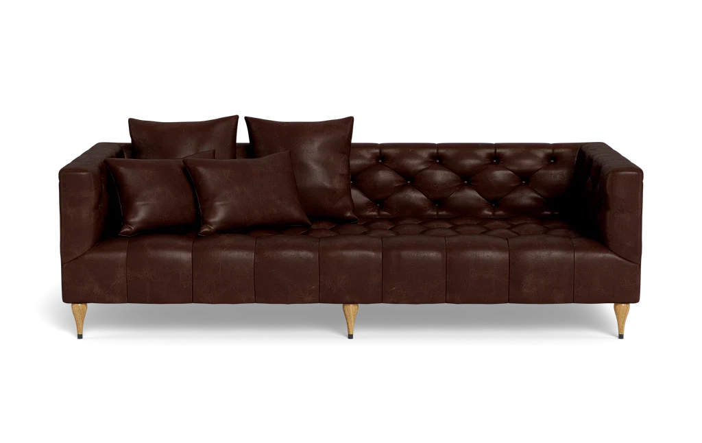 The Ms. Chesterfield Leather Custom Sofa, featuring a luxurious brown leather tufted design and wooden legs, is positioned against a plain background. It is elegantly adorned with four matching cushions, presenting a sleek and modern look.