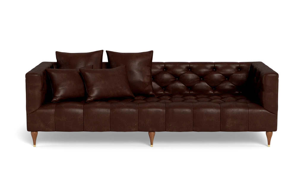 The Ms. Chesterfield Leather Custom Sofa is a dark brown leather piece featuring tufted cushions and paired with four matching pillows. It stands on short wooden legs and is showcased against a plain black background.