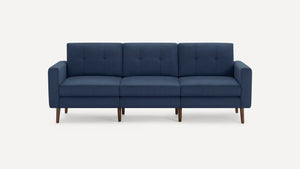 The Nomad Fabric Sofa is a modern, three-seater dark blue piece with a minimalist design, featuring wooden legs. It has tufted back cushions and straight armrests and is displayed against a plain white background.