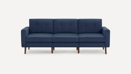 The Nomad Fabric Sofa is a modern, three-seater dark blue piece with a minimalist design, featuring wooden legs. It has tufted back cushions and straight armrests and is displayed against a plain white background.