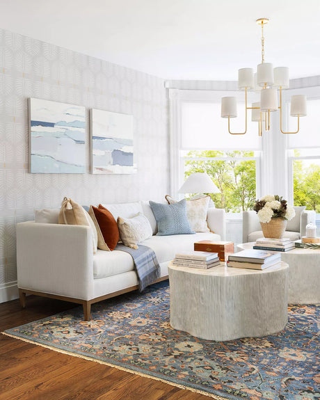 A modern living room features the Barton Sofa, adorned with various cushions and a throw blanket. Two abstract paintings hang on a patterned wall, and a round coffee table rests on a colorful rug, accompanied by a vase of flowers. Large windows allow natural light to fill the space.
