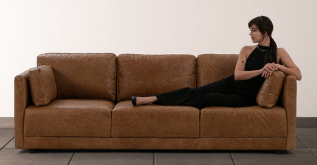 A person in dark clothing reclines on the Domus 93" Leather Sofa in chestnut, resting their head on a pillow and looking to the side. The background is a plain, light-colored wall.