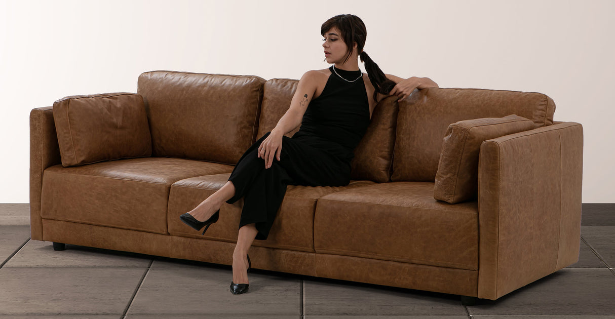 A person wearing a black dress and heels is sitting elegantly on the Domus 93" Leather Sofa in Chestnut, looking to the side. The background features a plain, light-colored wall.