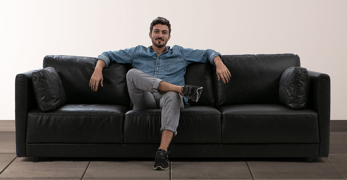 A person in a denim shirt and gray pants is lounging comfortably on the Domus 93" Leather Sofa in chestnut, accented with throw pillows and set against a simple background. They have their arms draped over the back of the sofa and legs crossed.