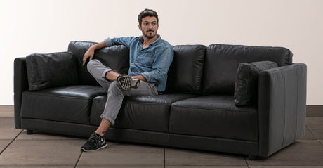 A man wearing a denim shirt and jeans is sitting cross-legged on a Domus 93" Leather Sofa in Chestnut. He is gazing off to the side, inside a room with a tile floor and a plain wall backdrop.