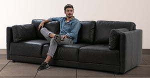 A man wearing a denim shirt and jeans is sitting cross-legged on a Domus 93" Leather Sofa in Chestnut. He is gazing off to the side, inside a room with a tile floor and a plain wall backdrop.