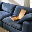 A dark blue sofa with a cardboard pizza box containing a few slices of pizza placed on the seat cushion. Sunlight filters through nearby curtains, creating a warm atmosphere in the room. A beige rug partially visible on the floor.