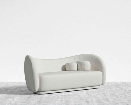 The Diane Sofa, featuring a contemporary light gray hue and a distinctive curved design, is positioned on a gray carpet against a simple white backdrop. Two matching rectangular cushions are placed on the seat.