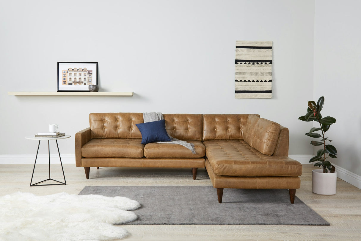 A contemporary living room showcasing the Eliot Leather Sectional with Bumper in brown, adorned with a gray throw and a blue pillow. A small side table nearby holds a cup. Completing the decor is a shelf displaying artwork, alongside a patterned wall hanging and a potted plant.