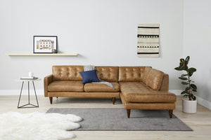 A contemporary living room showcasing the Eliot Leather Sectional with Bumper in brown, adorned with a gray throw and a blue pillow. A small side table nearby holds a cup. Completing the decor is a shelf displaying artwork, alongside a patterned wall hanging and a potted plant.