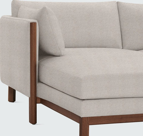 The Emmy Sectional is a modern corner sofa featuring gray upholstery and wooden legs, with a minimalist design characterized by clean lines and a cushioned seat.