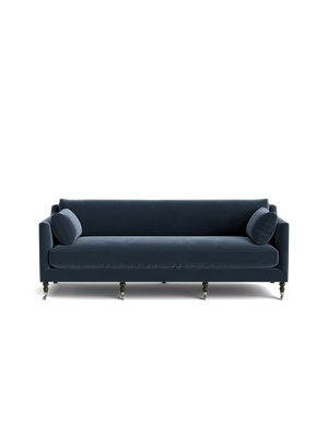 The Fabienne Sofa, in a dark blue hue, features a modern design with clean lines and cylindrical cushions set against a black backdrop. Its four visible legs add an elegant touch to its simple yet stylish appearance.