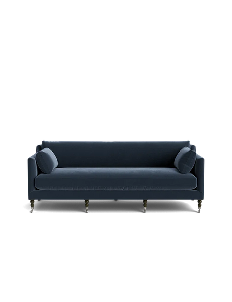 The Fabienne Sofa, in a dark blue hue, features a modern design with clean lines and cylindrical cushions set against a black backdrop. Its four visible legs add an elegant touch to its simple yet stylish appearance.