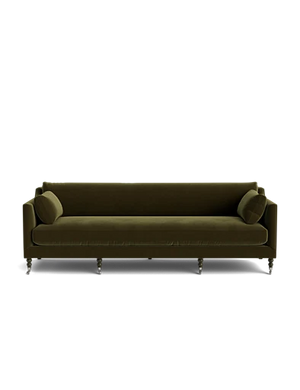 The Fabienne Sofa is upholstered in dark green velvet, featuring rounded armrests and accompanied by two matching cylindrical cushions. Its sleek metallic legs add a touch of modern elegance and comfort against a plain black background.